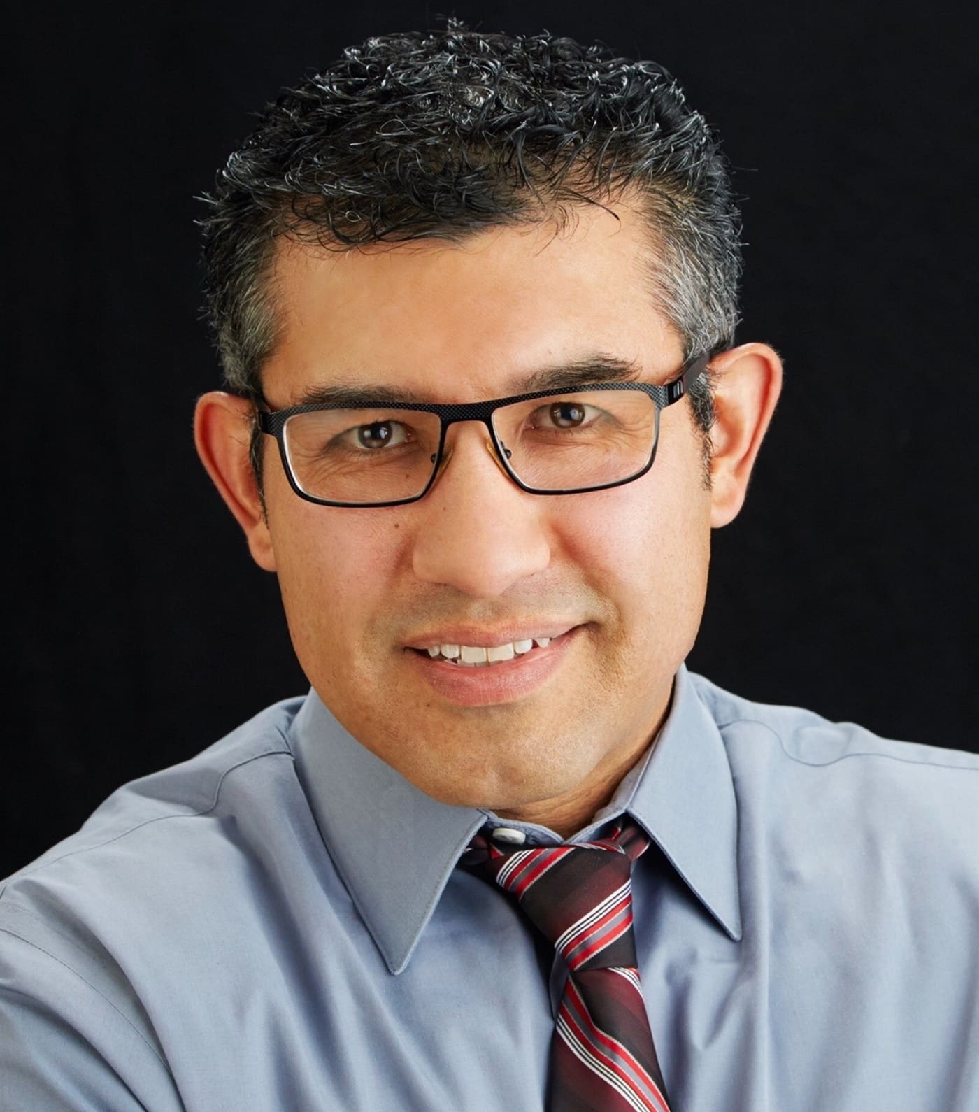 Headshot of Eli. He is a hespanic man with black and white short cropped hair. He wears black glasses and a tie.